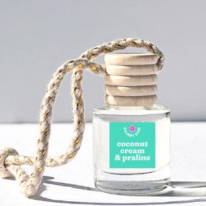 May include: A clear glass bottle with a wooden cap and a string for hanging. The bottle has a turquoise label with white text that reads "coconut cream & praline". The label also has a pink smiley face with the text "chubby star" above it.