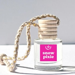 May include: A small, clear glass bottle with a wooden cap and a woven cord for hanging. The bottle has a bright pink label with the words "snow pixie" and a star logo. The cord is beige with gold accents.