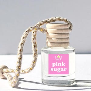 May include: A clear glass bottle with a pink label that says "pink sugar" and a yellow smiley face with a star. The bottle has a wooden cap and is attached to a white and gold rope.