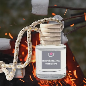 May include: A clear glass car air freshener with a wooden cap and a rope hanger. The label reads "marshmallow campfire" with a pink smiley face logo. The background shows marshmallows roasting over a campfire.