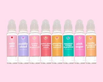 May include: A row of eight roll-on perfume oil bottles with colorful labels and clear glass. Each bottle has a different scent name, including "Only You," "Cuddly Cloud," and "Pink Sugar." The background is a solid light pink.