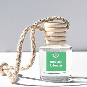 May include: A clear glass bottle with a green label that says "Chubby Star cactus bloom". The bottle has a wooden cap and is attached to a white string with gold accents.
