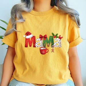 Könnte beinhalten: Senfgelbes T-Shirt mit dem Wort "Mama" in einem weihnachtlichen Design. Die Buchstaben sind mit einem Weihnachtsmannhut, einer Schleife, Stechpalmen und Tupfen verziert. Eine Tasse heiße Schokolade und ein Lebkuchenmann sind ebenfalls abgebildet.