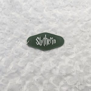 May include: A green keychain tag with the word "Slytherin" in white lettering. The tag has a retro hotel key design and a small metal ring. The background is a white, textured surface.