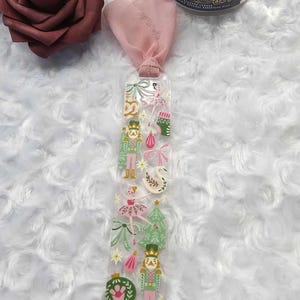 Könnte beinhalten: Ein klarer Acryl-Lesezeichen mit rosa Band, das farbenfrohe Illustrationen von Nussknackern, Ballerinas, Weihnachtsbäumen und anderen festlichen Designs zeigt. Der Lesezeichen ist rechteckig und hat eine glatte Oberfläche.