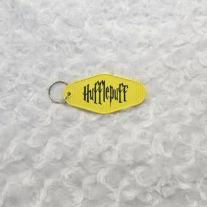 May include: A yellow keychain with the word "Hufflepuff" in black, set against a white, textured background. The keychain has a retro motel style design with a metal ring for attaching keys.