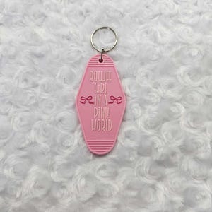 May include: A pink motel-style keychain with the phrase "Boujie Girl in a Pink World" in white lettering. The keychain has a silver ring and two bow graphics. The background is a white, fluffy surface.