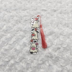 Könnte beinhalten: Ein klarer Acryl-Lesezeichen mit drei Cartoon-Geistern, die Bücher lesen. Jeder Geist ist weiß mit schwarzen Augen und ist mit rosa Blumen und einer rosa Quaste verziert. Das Lesezeichen ist etwa 15 cm lang.