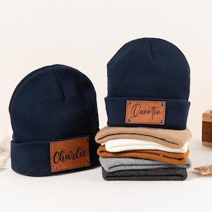 Può includere: Due berretti blu navy con targhette con nome in pelle marrone. Un berretto ha il nome "Charlie" e l'altro il nome "Quentin". Una pila di berretti piegati in vari colori è accanto ai berretti. Una macchina fotografica di legno e oggetti decorativi sono sullo sfondo.