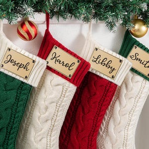 May include: Four cable-knit Christmas stockings featuring a ribbed cuff and a rectangular wooden name tag attached with four metal rivets. The stockings are colored in green, white, red, and white, with the names Joseph, Karol, Libby, and Smith.