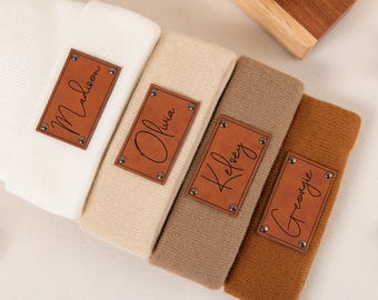 May include: Four folded knit beanies in cream, beige, brown, and rust colours. Each beanie has a rectangular brown leather patch with a name in cursive script: Madison, Olivia, Kelsey, and Georgie. The patches are secured with small silver rivets.