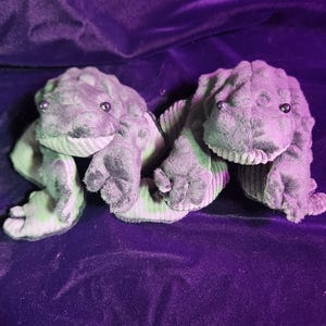 May include: Two plush green frog toys with black eyes. The frogs are lying on a purple velvet surface.