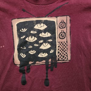 以下が含まれることがあります： ビンテージテレビの画面のグラフィックが描かれたマルーンのTシャツ。画面は白黒の目でいっぱいになっており、テレビから黒い塗料が垂れている。