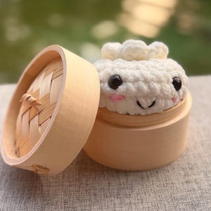 Könnte beinhalten: Ein weißer, gehäkelter Plüsch-Dumpling mit schwarzen Kunststoffaugen, einem gestickten Mund und rosa Wangen, der in einem runden, hellen Bambusdampfgarer sitzt, wobei der passende Deckel neben dem Behälter platziert ist.