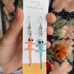 May include: A pair of beaded lizard earrings on a white card with the text "mig makes". One earring is light blue and the other is peach.