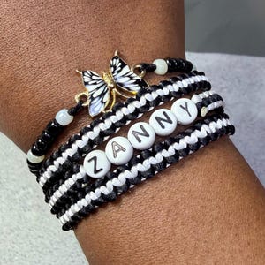 May include: A multi-strand bracelet featuring a gold-toned butterfly charm with black and white enamel wings. The design includes black and white woven cord bands, small black and white beads, and white round letter beads spelling out ZANNY.