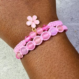 May include: A pink bracelet featuring a woven cord design with translucent pink spherical beads and faceted pink crystal accents. A small, five-petaled pink enamel flower charm with a gold-toned center is attached to the bracelet.