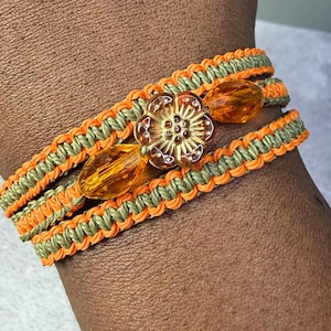 May include: A multi-strand bracelet featuring woven sage green and bright orange cord. The design includes a central flower-shaped amber-colored bead flanked by two faceted, translucent amber-colored beads, creating a layered, textured appearance.