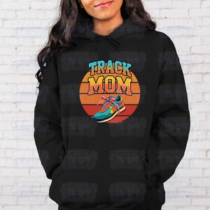 Peut inclure: Sweat &agrave; capuche noir avec un graphisme color&eacute;. Le graphisme pr&eacute;sente les mots "TRACK MOM" en bleu et orange, avec un design de chaussure de course sur fond de coucher de soleil.