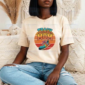 Peut inclure: Un t-shirt beige avec l'inscription "TRACK MOM" dans un design r&eacute;tro color&eacute;. Le motif comprend une chaussure de course dans un cercle de coucher de soleil. Le t-shirt est en mati&egrave;re douce, adapt&eacute; au port d&eacute;contract&eacute;.