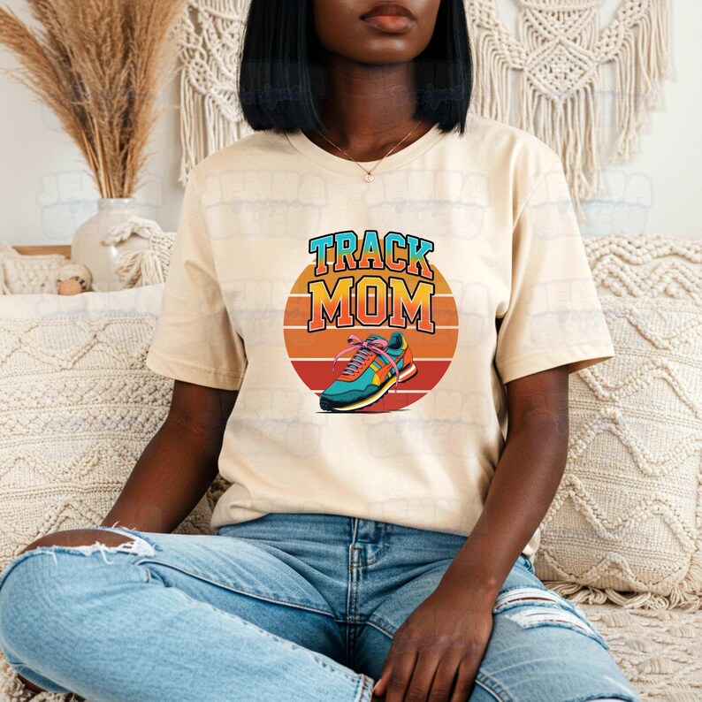 Peut inclure: Un t-shirt beige avec l'inscription "TRACK MOM" dans un design r&eacute;tro color&eacute;. Le motif comprend une chaussure de course dans un cercle de coucher de soleil. Le t-shirt est en mati&egrave;re douce, adapt&eacute; au port d&eacute;contract&eacute;.