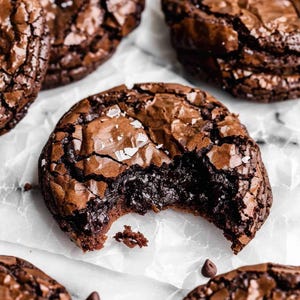 Peut inclure: Gros plan sur plusieurs cookies au chocolat noir avec une surface craquelée et texturée. Un cookie a une bouchée, révélant un intérieur moelleux et fondant. Les cookies sont saupoudrés de gros sel et de pépites de chocolat.