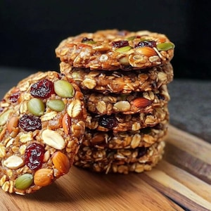 Puede incluir: Una pila de galletas de granola redondas y texturizadas con avena, nueces, semillas y arándanos rojos secos visibles. Las galletas están dispuestas sobre una tabla de cortar de madera, con un fondo oscuro. Las galletas son marrones, rojas, verdes y beige.
