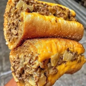 May include: Two halves of a Philly cheesesteak sandwich. The sandwiches have a golden-brown crust, filled with chopped meat, melted cheese, and onions. The cheese is a bright yellow colour.