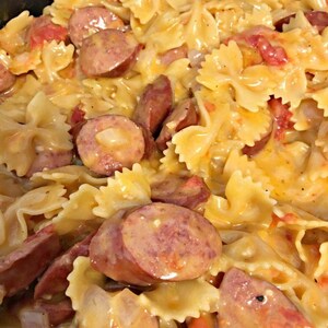 Peut inclure: Gros plan sur un plat de pâtes avec des farfalles, des saucisses tranchées et des tomates coupées en dés dans une sauce crémeuse. Le plat est un mélange de couleurs jaune, rouge et marron.