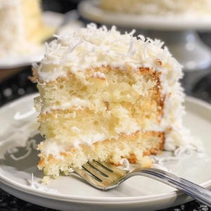 Peut inclure: Une part de gâteau à la noix de coco sur une assiette blanche avec une fourchette. Le gâteau a plusieurs couches de génoise jaune avec du glaçage blanc et de la noix de coco râpée sur le dessus.