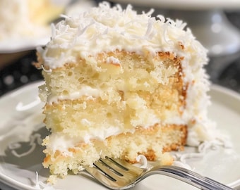 May include: A slice of coconut cake on a white plate with a fork. The cake has multiple layers of yellow cake with white frosting and shredded coconut on top. Another cake is visible in the background.