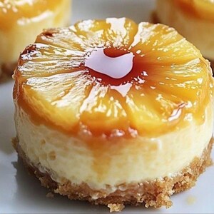 Può includere: Primo piano di una mini cheesecake all'ananas rovesciata. Il dessert presenta una base dorata, un ripieno cremoso bianco e una copertura di fette di ananas caramellate. Una glassa lucida ricopre l'ananas.