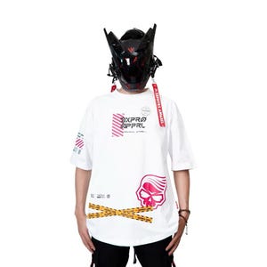 Pode incluir: Camiseta branca com gráficos rosa e vermelhos, incluindo uma caveira e o texto "DXPRO APPRL". A camiseta tem mangas curtas e uma estética streetwear. Detalhes em fita adesiva amarela.