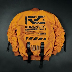 May include: Mustard-coloured bomber jacket with black graphic text and design elements. Features include a front pocket with black straps and buckles, and ribbed cuffs and collar. The jacket has the text "Krmln Genesis System" and "Type Future 99".