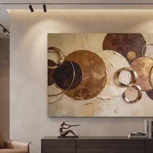 Peut inclure: Grande œuvre d'art abstrait mural avec des cercles superposés dans des tons de marron, d'or et de crème. Des anneaux métalliques sont intégrés au design. L'œuvre est exposée au-dessus d'un meuble en bois foncé avec des objets décoratifs.