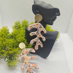 May include: A pair of dangle earrings with a floral pattern on a white background. The earrings are made of polymer clay and have a gold-colored metal top.