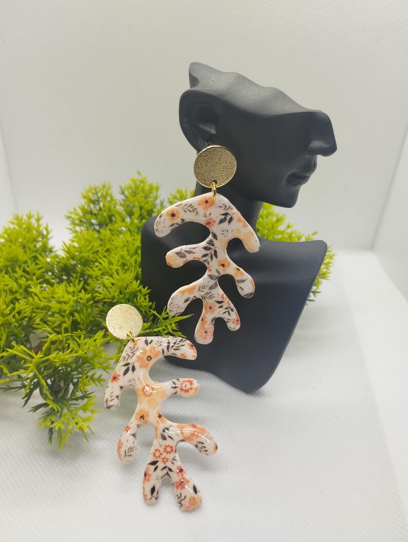 May include: A pair of dangle earrings with a floral pattern on a white background. The earrings are made of polymer clay and have a gold-colored metal top.