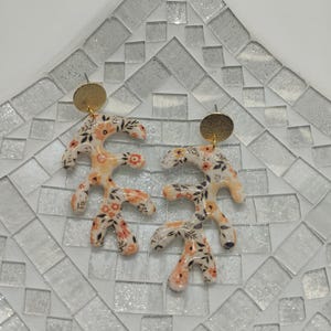 May include: A pair of floral patterned earrings with gold accents. The earrings are shaped like branches with small flowers and leaves.