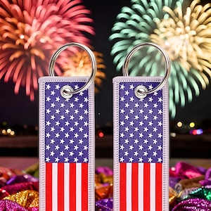 May include: Two keychains with silver rings and a purple fabric design featuring a white star pattern and red and white stripes. The keychains are in front of a blurred background of fireworks.