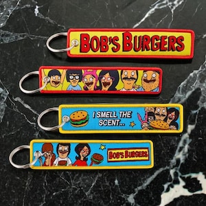 May include: Four colorful keychains featuring characters from the animated TV show Bob's Burgers. The keychains are made of fabric and have a yellow, red, and blue color scheme. The keychains feature the text "Bob's Burgers", "I smell the scent...", and "Bob's Burgers".
