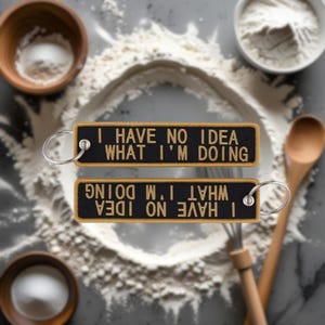 May include: Two black keychains with the text "I HAVE NO IDEA WHAT I'M DOING" in gold lettering. One keychain is right-side up, and the other is upside down. The keychains are on a surface with flour and baking tools.