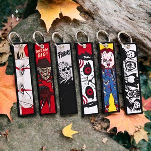 May include: Six colorful keychains featuring horror movie characters. The keychains are black and white with red, blue, and yellow accents. The characters include Freddy Krueger, Jason Voorhees, Chucky, and Michael Myers. The keychains are made of fabric and have a metal ring for attaching to keys.