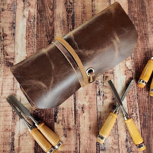 May include: A brown leather roll-up case with a strap and buckle closure. The case is open and contains four wooden-handled chisels with metal blades.