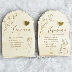 Könnte beinhalten: Zwei Holzschilder mit gewölbten Oberteilen. Eines sagt "Nounou" und das andere "Maîtresse" in Kursivschrift. Beide haben ein goldenes Herz, Blumendesigns und französischen Text. Die Schilder sind hellbraun mit gravierten Details.