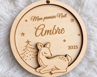 Peut inclure: Une décoration de Noël ronde en bois gravée avec le texte Mon premier Noël, le prénom Ambre et l'année 2025. Le motif comprend une figurine de faon en bois en relief, un petit sapin de Noël et plusieurs étoiles.