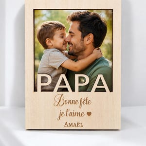 以下が含まれることがあります： 父と子供が抱き合っている写真が入った木製のフォトフレーム。「PAPA」の文字が上部に切り抜かれ、「Bonne f&ecirc;te je t'aime」とハートのマークが下にあります。名前「AMA&Euml;L」が一番下にあります。