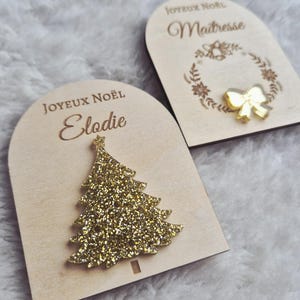 Peut inclure: Deux ornements de Noël en bois. L'un présente un sapin de Noël en paillettes dorées, l'autre un nœud doré. Les deux portent les mots "JOYEUX NOËL" et un nom ou un titre gravé. Ils sont sur une surface blanche et douce.
