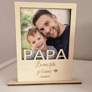 以下が含まれることがあります： 父と子の写真が入った、明るい茶色の木製フォトフレーム。「PAPA」の文字が上部に切り抜かれ、「Bonne f&ecirc;te je t'aime」と「LEROY」の名前が刻まれています。