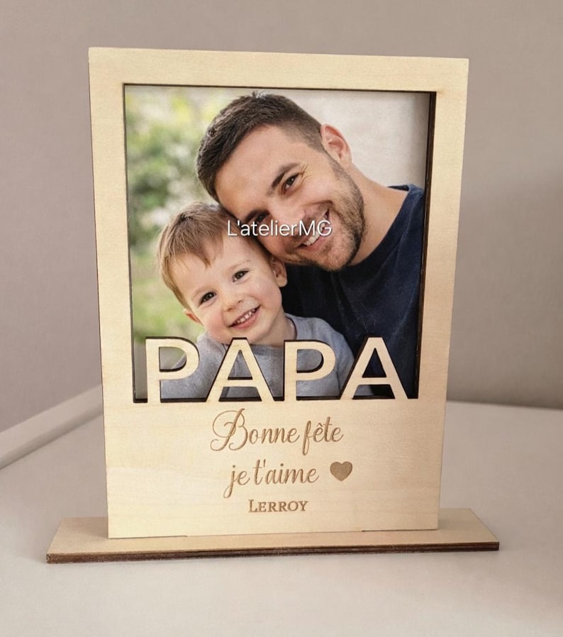 以下が含まれることがあります： 父と子の写真が入った、明るい茶色の木製フォトフレーム。「PAPA」の文字が上部に切り抜かれ、「Bonne f&ecirc;te je t'aime」と「LEROY」の名前が刻まれています。