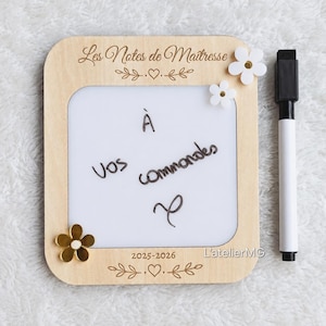 Peut inclure: Tableau blanc en bois avec le texte "Les Notes de Maîtresse" et "À Vos Commandes". Le tableau blanc comporte des décorations florales et un marqueur noir. Le texte "2025-2026" est également visible.
